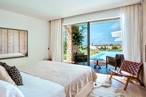 A hotel room with a bed, a small table, a chair, and a view of a pool and landscape. - Villa Isidor with private pool - My Rental Homes (San Jose)