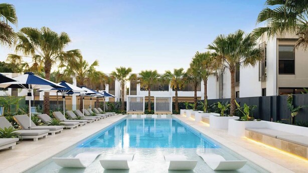 A swimming pool with lounge chairs and umbrellas, surrounded by palm trees and modern buildings. - The Capreae - LUX Townhouse Surfers Paradise Pool (Surfers Paradise)