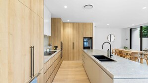A modern kitchen with wooden cabinets, a central island, and a dining area with wooden chairs. - The Capreae - LUX Townhouse Surfers Paradise Pool (Surfers Paradise)