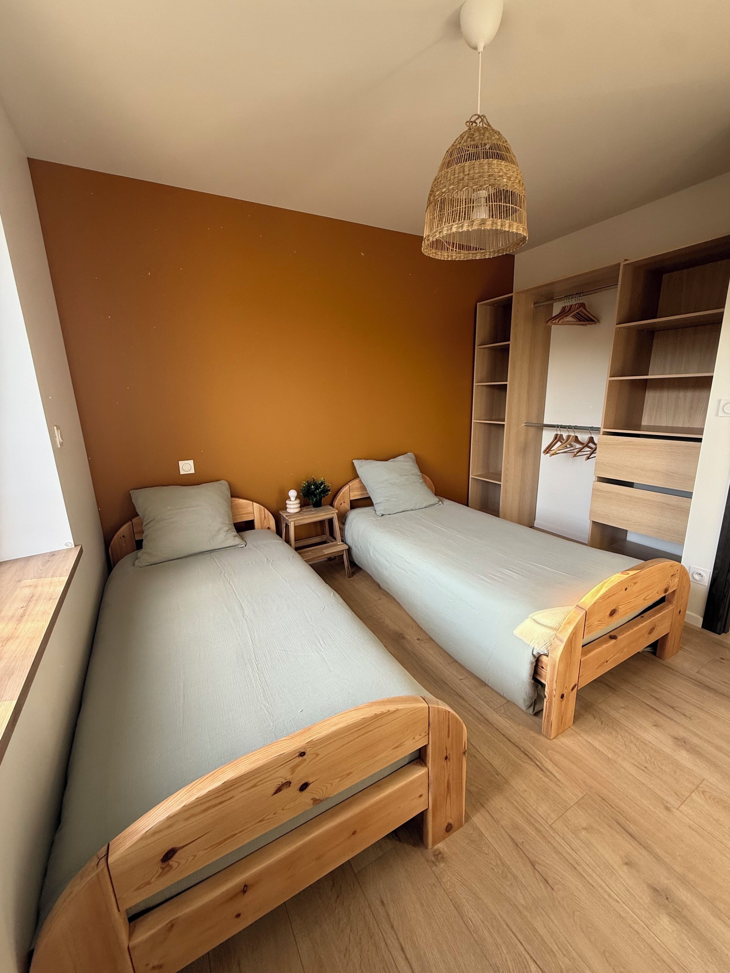 Un dormitorio con dos camas de madera, una lámpara de techo de fibra natural, una mesita de noche pequeña y un armario con estantes abiertos.