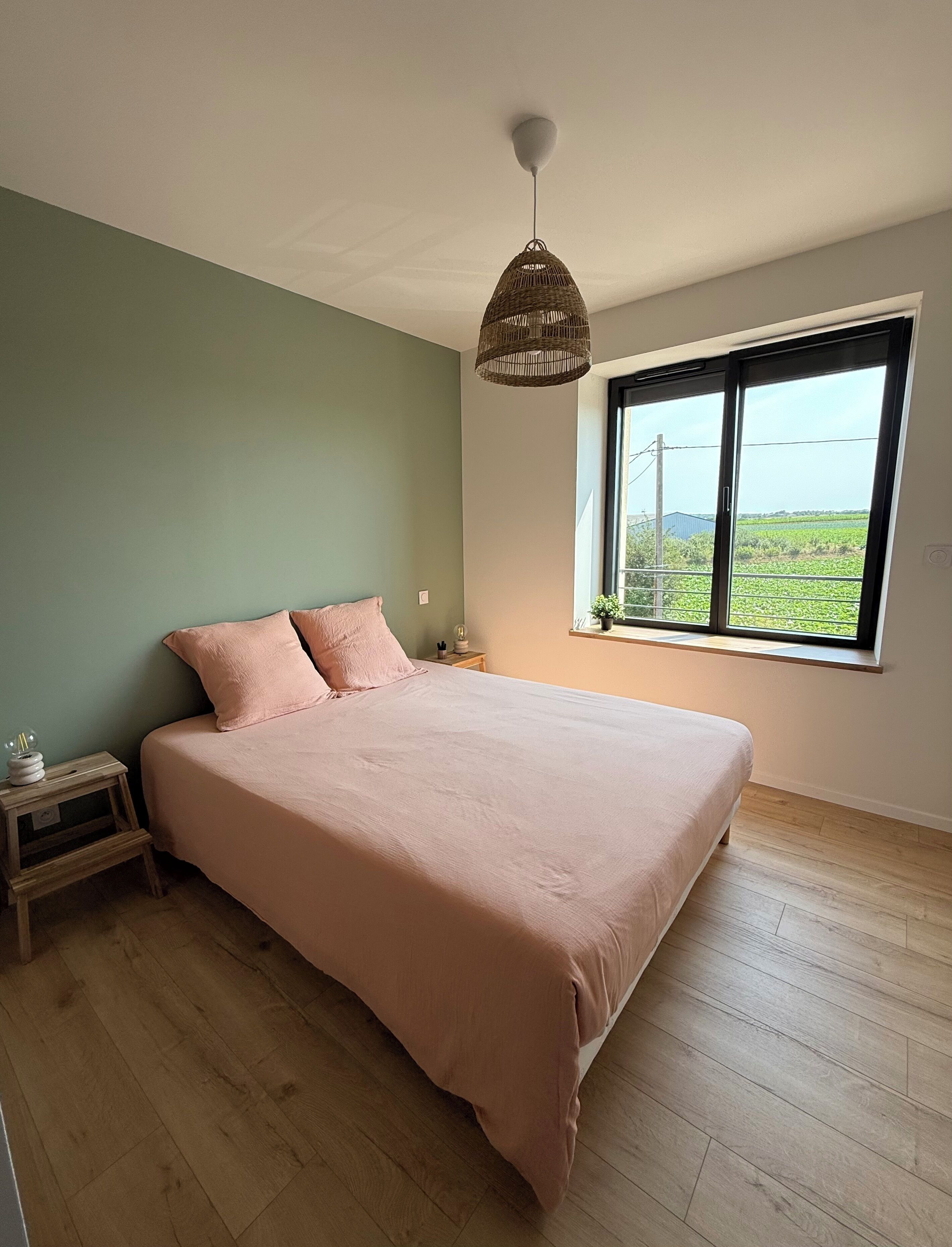 A bedroom with a large bed, a bedside table, a nightstand, a hanging light fixture, and a window with a view of greenery.