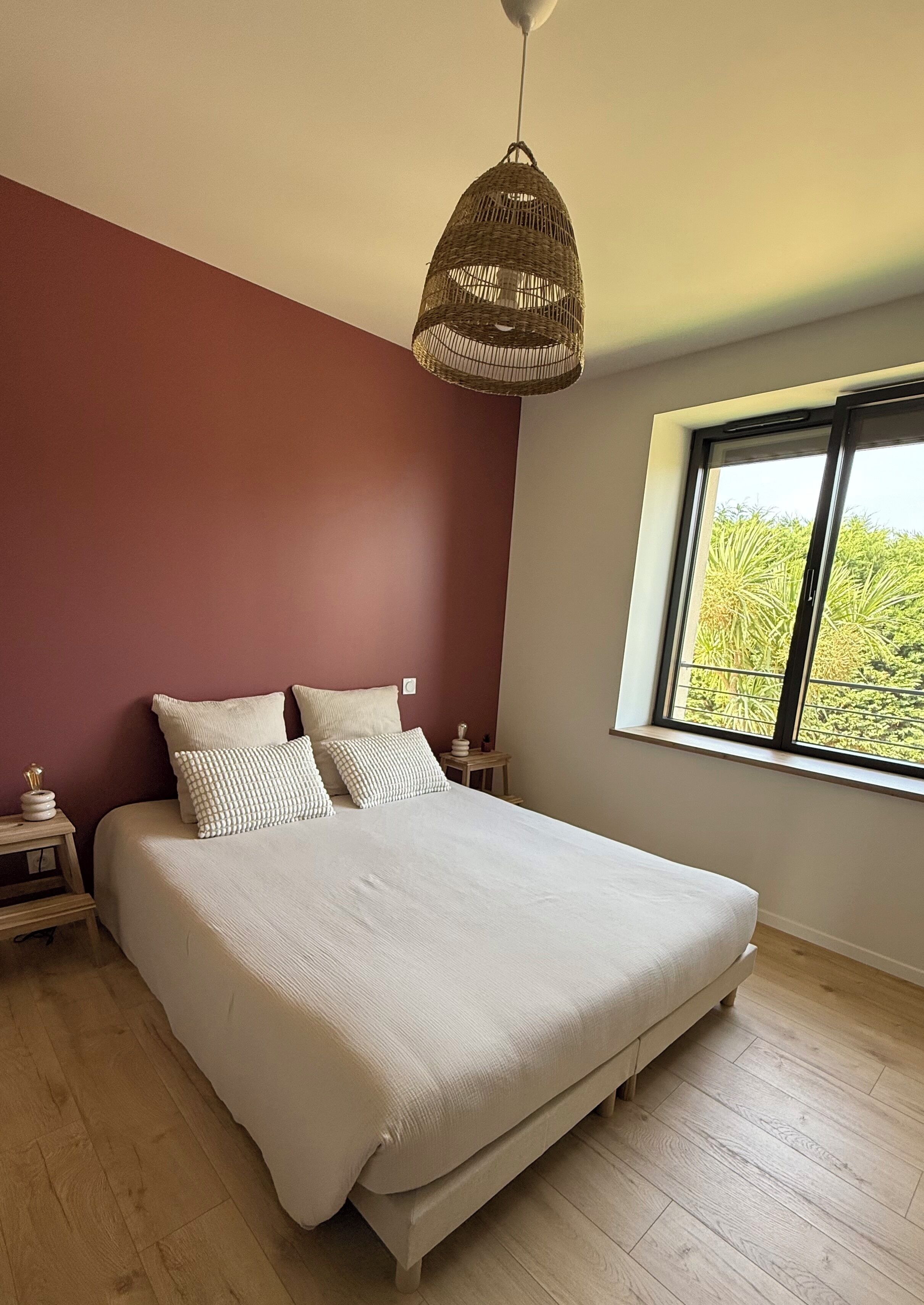 Une chambre avec un grand lit, une table de chevet, une commode, un luminaire suspendu et une fenêtre donnant sur un paysage verdoyant.