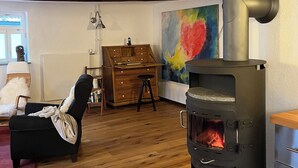 Un salon chaleureux avec un plancher en bois, un fauteuil noir, une commode en bois, un tableau coloré et un poêle à bois.
