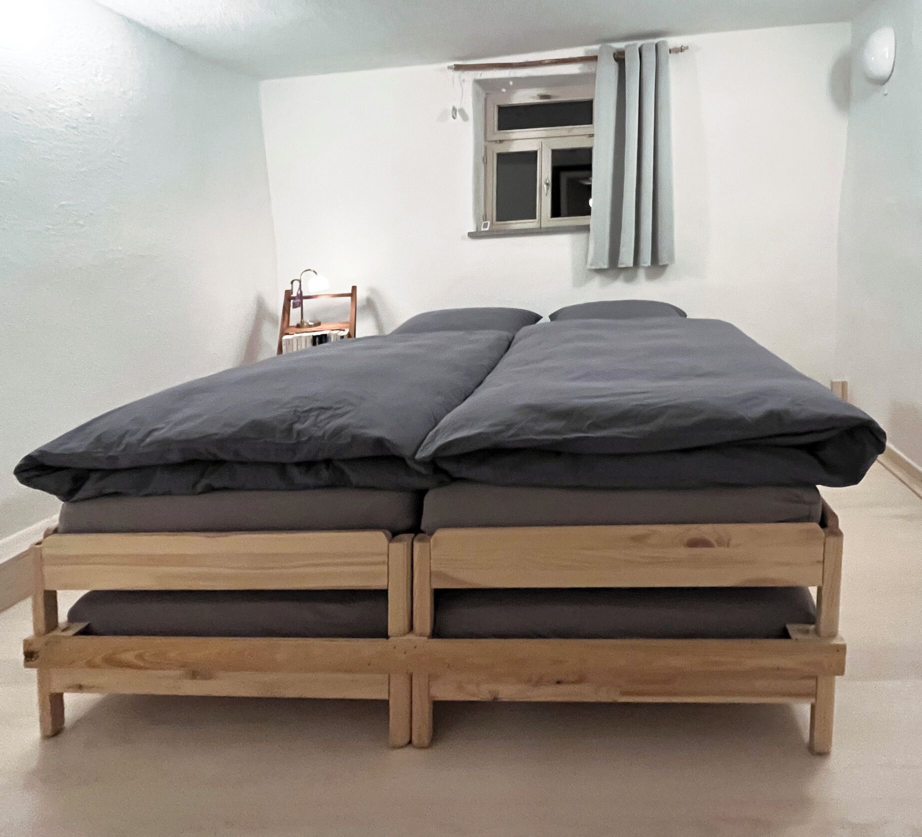 Un lit simple avec un cadre en bois et une literie sombre, dans une pièce aux murs blancs et dotée d’une fenêtre avec des rideaux.