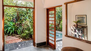 Un pasillo con una puerta de madera, suelo de baldosas y un espejo que refleja una planta y una estantería.