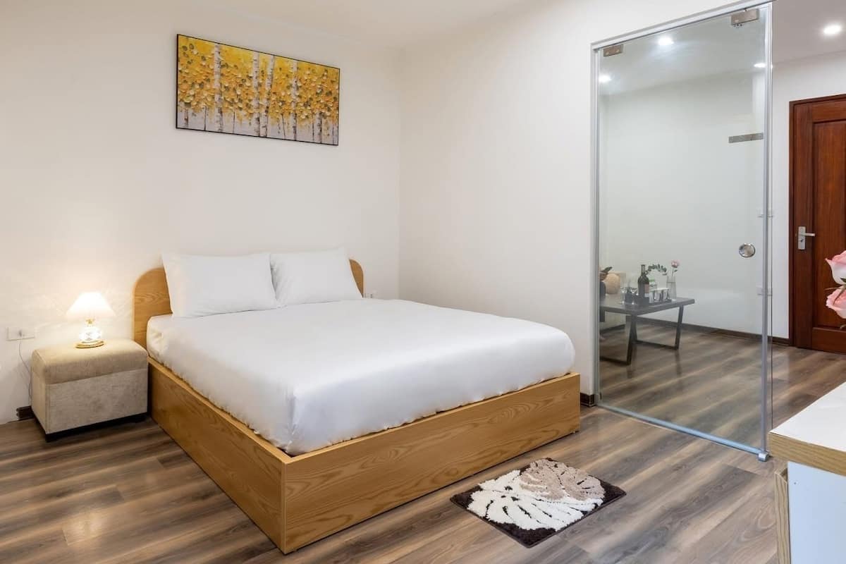 A bedroom with a bed, bedside table, lamp, artwork, and a glass partition leading to a bathroom.