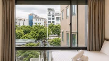 Habitación de hotel con un amplio ventanal corredero, cama con ropa de cama blanca y vista a edificios y árboles.