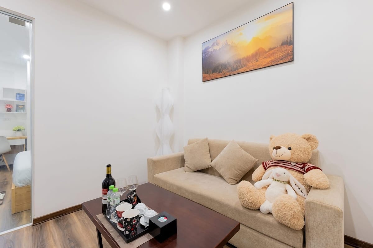 A cozy room with a beige sofa, a teddy bear, a white bunny, a wine bottle, glasses, and a tray with cups and a box.