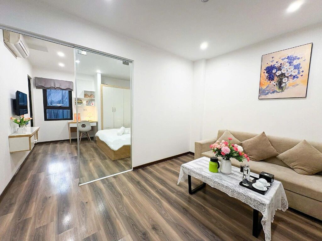 A modern living room with a sofa, a coffee table with decorative items, a TV, and a glass partition leading to a bedroom.