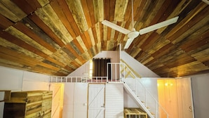 An attic room with a sloped ceiling, wooden beams, a white staircase, and a ceiling fan. - PILIBHIT Forest & Feast (Puranpur)