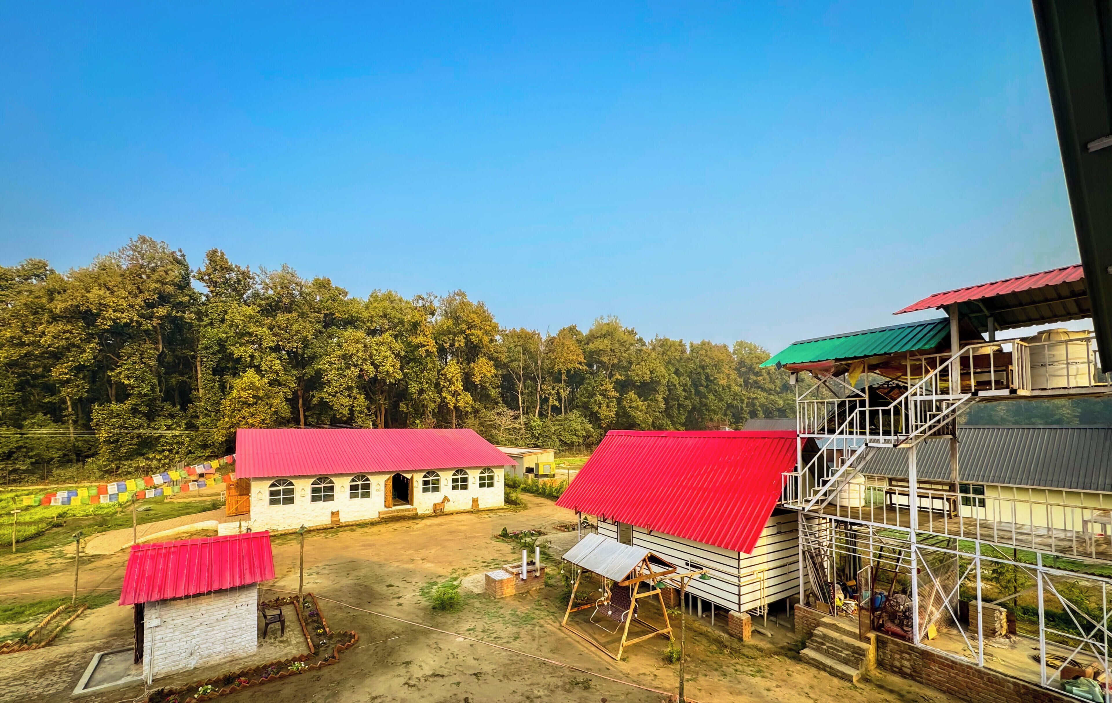 Entorno rural con un edificio blanco de techo rojo, un edificio más grande con techo verde y una estructura en construcción con andamios.