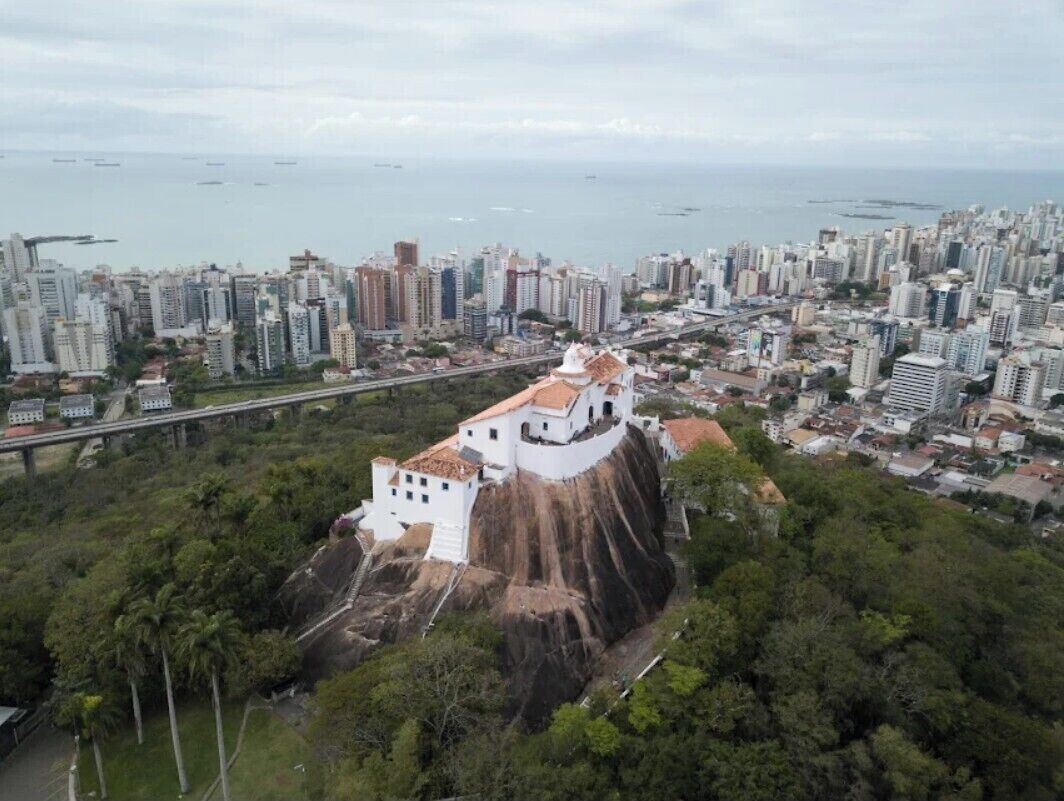 Un paysage urbain côtier avec un imposant bâtiment blanc perché sur une falaise, entouré d’une végétation luxuriante et d’un quartier animé avec de nombreux immeubles de grande hauteur.