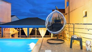Un espace piscine agrémenté d’un fauteuil suspendu, d’une table avec des livres et de guirlandes lumineuses.
