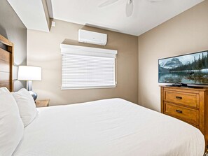 Une chambre à coucher comprenant un lit, un téléviseur, un ventilateur de plafond, une fenêtre avec des stores et un climatiseur fixé au mur.
