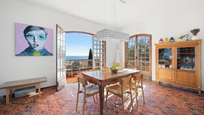A dining area with a table and chairs, a bench, a large window with a view, and a cabinet with glass doors. - Maison Soleillade – A rustic, peaceful home with a view of the sea (Roquebrune-Cap-Martin)