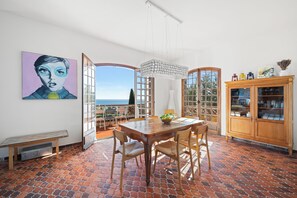 A dining area with a table and chairs, a bench, a large window with a view, and a cabinet with glass doors. - Maison Soleillade – A rustic, peaceful home with a view of the sea (Roquebrune-Cap-Martin)