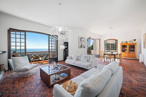 A spacious living area with a large window overlooking the sea, a white sofa, a coffee table, and a dining area with a wooden cabinet. - Maison Soleillade – A rustic, peaceful home with a view of the sea (Roquebrune-Cap-Martin)