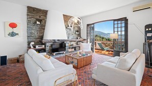 A living room with a stone fireplace, white sofas, a coffee table, and a view of the outdoors. - Maison Soleillade – A rustic, peaceful home with a view of the sea (Roquebrune-Cap-Martin)
