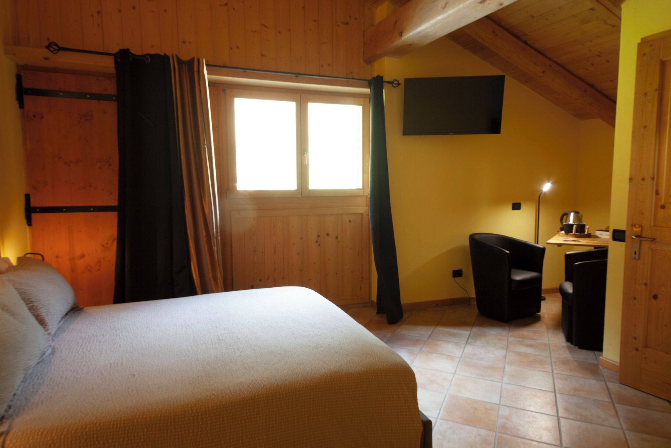 A room with a bed, a window with curtains, a television on the wall, and a small table with a lamp.