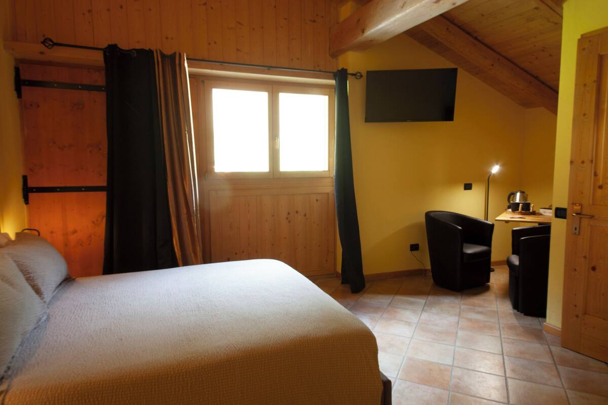 Una stanza con un letto, una finestra con tende, una televisione appesa al muro e un tavolino con una lampada.