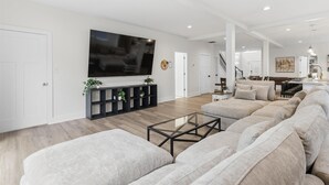 Een moderne woonkamer met een grote flatscreen televisie, een hoekbank, een glazen salontafel en een zwarte entertainmentkast met planten.