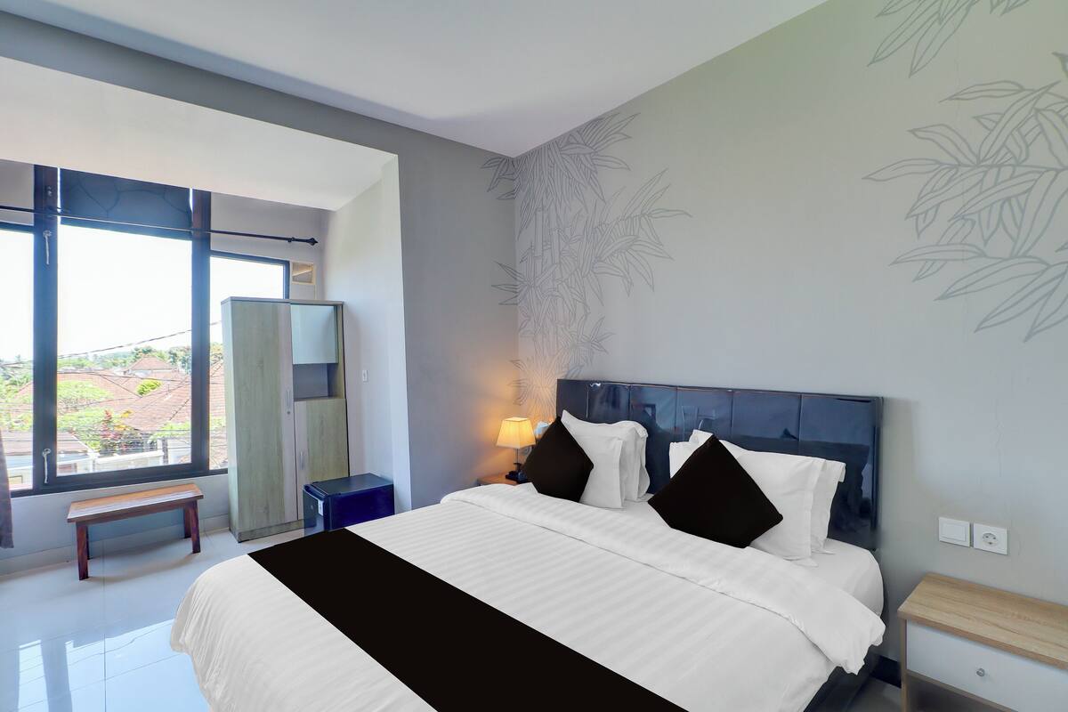 Une chambre d’hôtel moderne, dotée d’un grand lit, d’une banquette, d’une table de chevet avec une lampe et offrant une vue sur l’extérieur grâce à une grande fenêtre.
