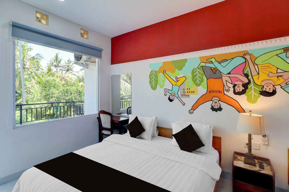 Une chambre d’hôtel comprenant un lit, un bureau, une chaise, une fenêtre donnant sur un espace verdoyant et une fresque murale.