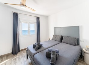A double bed with grey linens and towels, a ceiling fan, a window with curtains, and a view of the sea. - Beachfront Holiday Home in Malaga (Mijas)