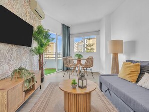 Ein modernes Wohnzimmer mit einer Couch, einem runden Couchtisch, einem an der Wand befestigten Fernseher und einem Balkon mit Blick auf Gebäude.