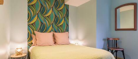 Un dormitorio con cabecera estampada, cama con manta texturizada, lámpara de noche y una silla.