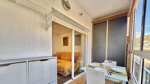 Een compacte kamer met een eettafel voor twee personen, een bank en een schuifglazen deur naar een balkon.