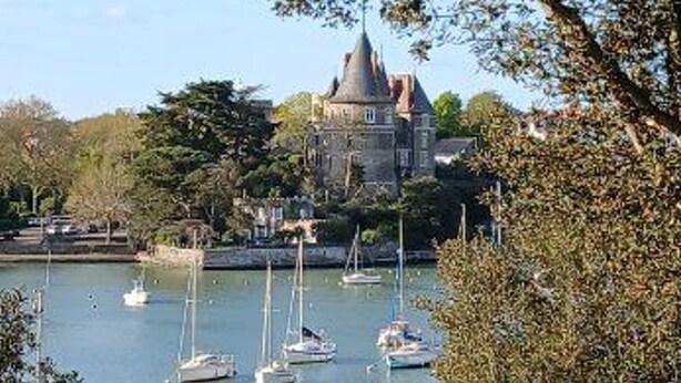Un bâtiment qui ressemble à un château, avec une tour, entouré d’une étendue d’eau où naviguent plusieurs voiliers.