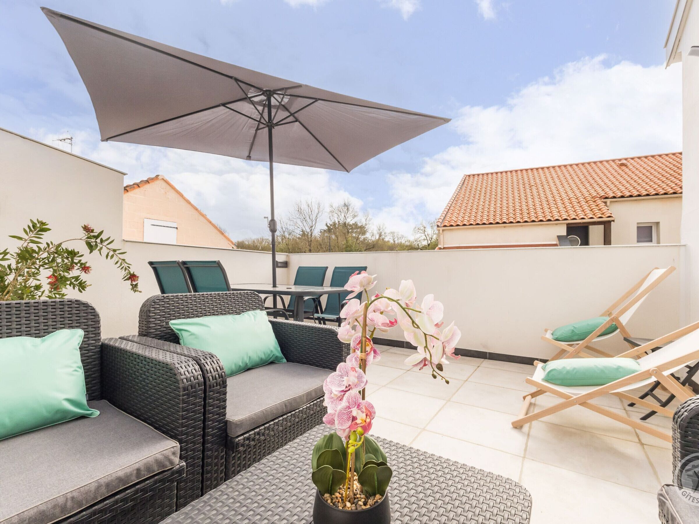 Une terrasse extérieure aménagée avec des meubles en osier, une table et des chaises, un grand parasol et un pot d’orchidée.