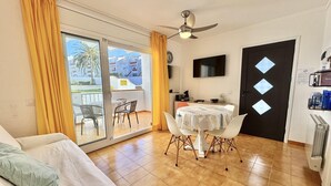 A living room with a sofa, a dining table with chairs, a ceiling fan, a TV, and a sliding glass door leading to an outdoor area. - Cozy Apartment with Pool and Terrace in Puig Rom, 2km from Roses Beach - Sleeps 4 (Roses)