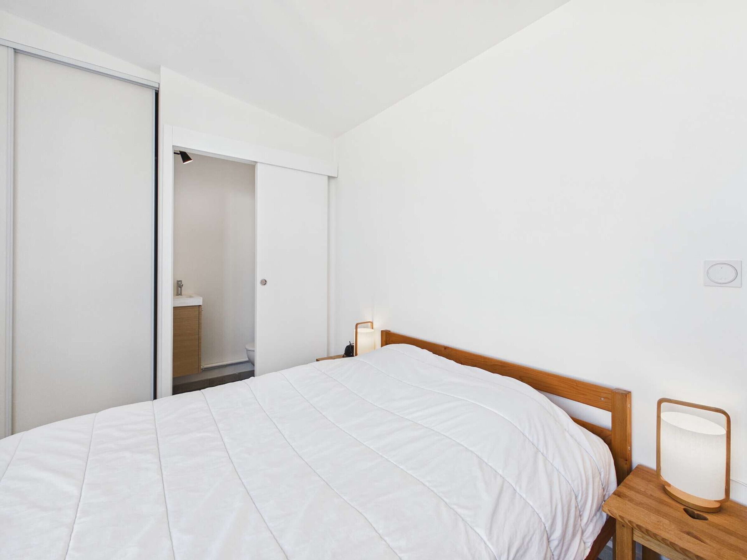 Ein Schlafzimmer mit einem Bett, einem Nachttisch aus Holz, einer Nachttischlampe, einem Kleiderschrank und einem durch eine offene Tür einsehbaren Badezimmer.