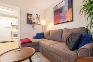 A living room with a sofa, a coffee table, and a lamp. There is a kitchen visible in the background.