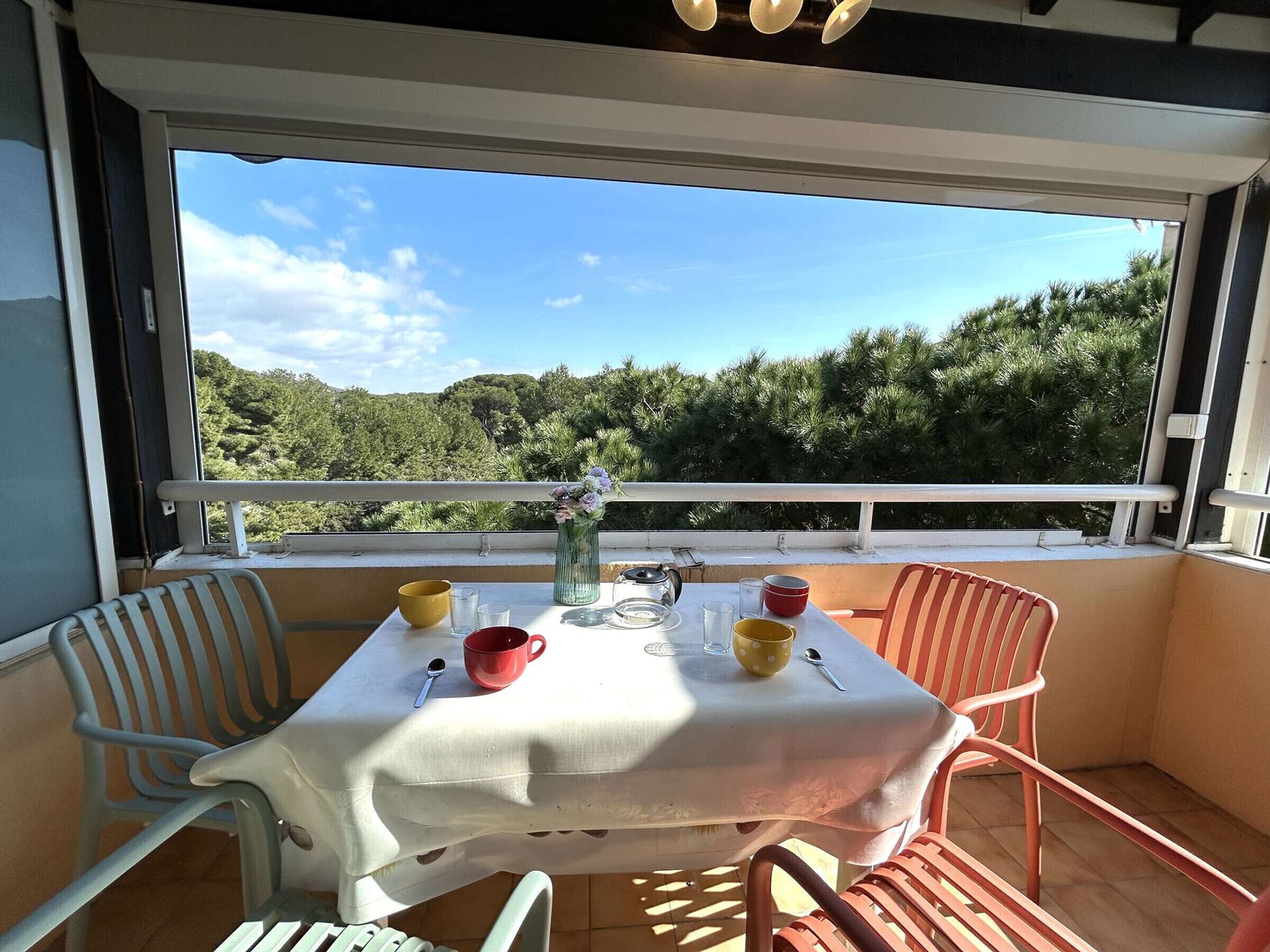 A balcony with a table set for two, chairs, a vase with flowers, and a view of a forested hillside.