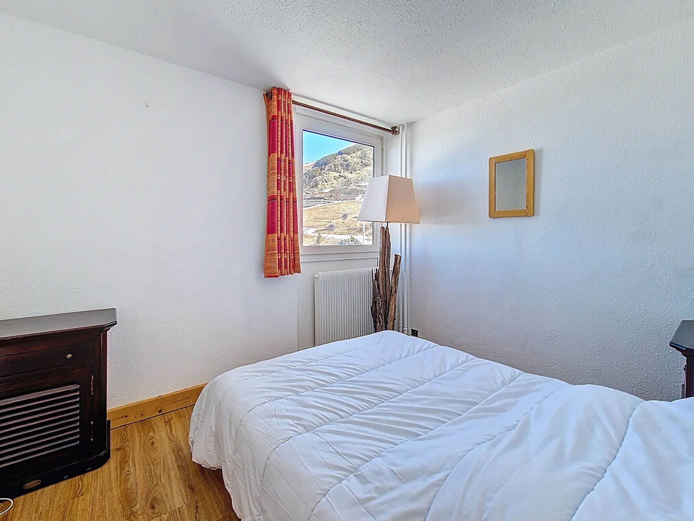 Een slaapkamer met een bed, een radiator, een lamp, een raam met gordijnen en een houten dressoir.