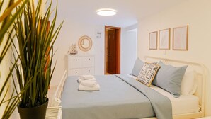 Un dormitorio con cama, mesita de noche, planta en maceta y una puerta que da a otra habitación.