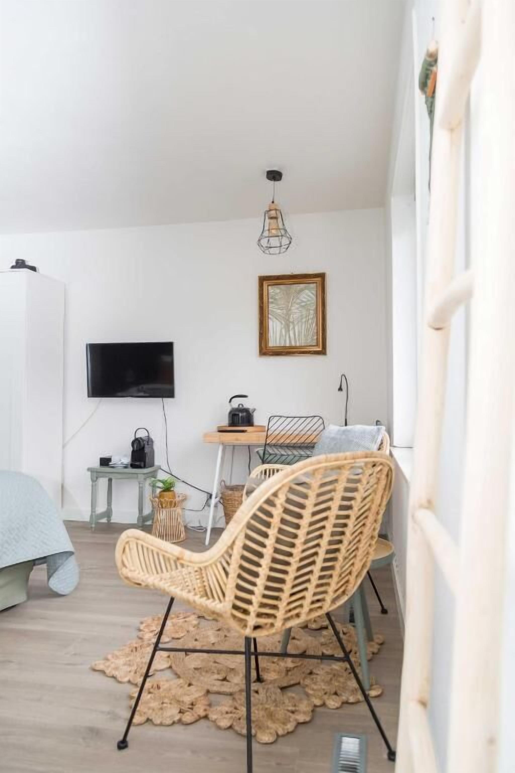 Una camera con un letto, una TV, un tavolo con un bollitore, una sedia in vimini e una lampada a soffitto.