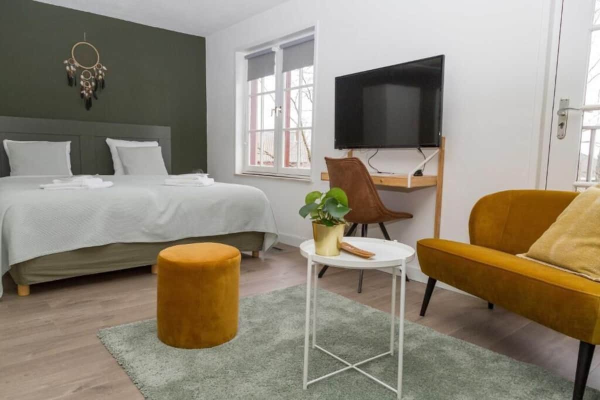 A bedroom with a bed, a small round ottoman, a desk with a chair, and a television.