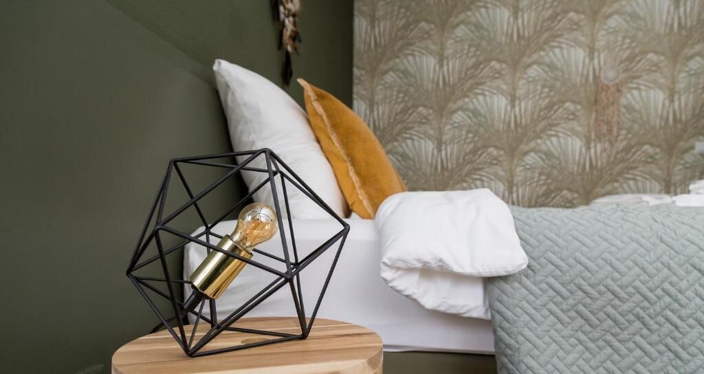 Una cama con una manta blanca, una lámpara geométrica sobre una mesita de noche de madera y un papel tapiz estampado de fondo.