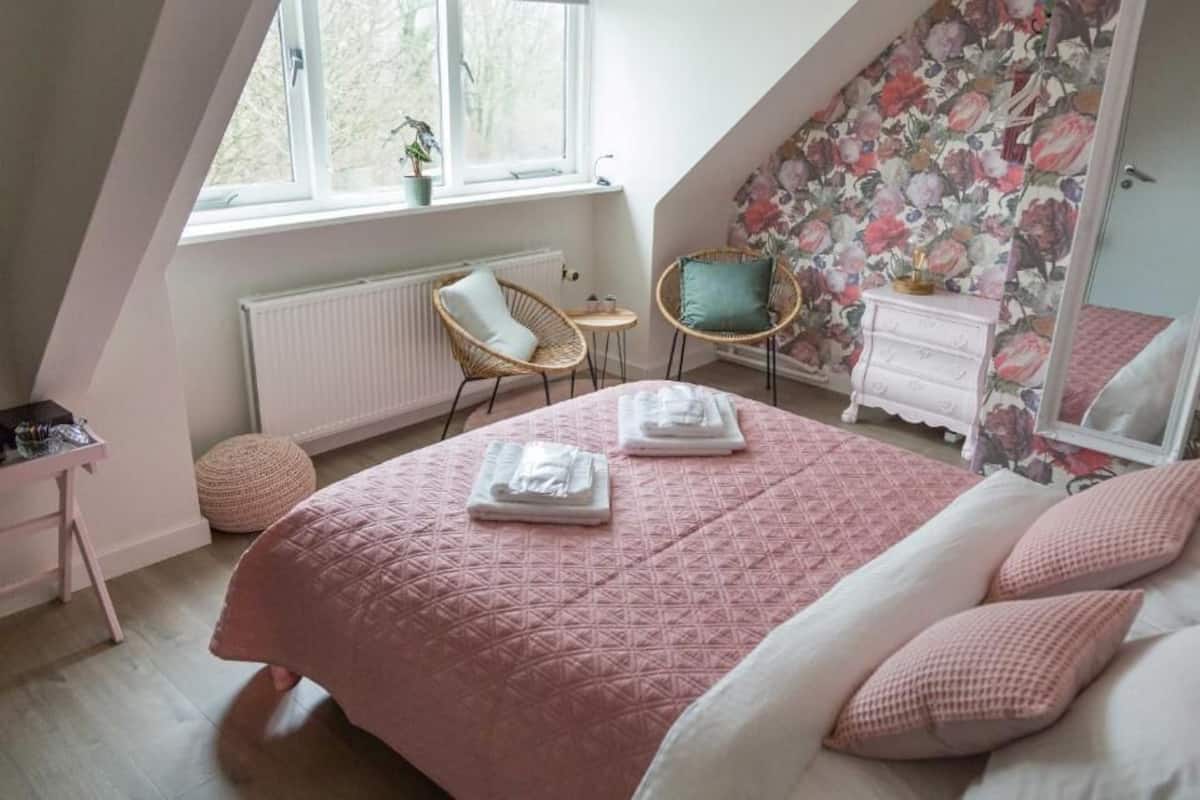 A bedroom with a pink quilted bedspread, a white dresser, a wicker chair, and a floral wallpaper.