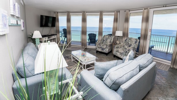 En moderne stue med sofa, lænestole, et sofabord og et fjernsyn. Der er gardiner og en balkon med udsigt til havet.
