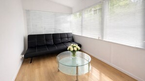 Een hoekzaal met een zwarte leren bank, een glazen tafel met een bloevas en grote ramen met zonwering.