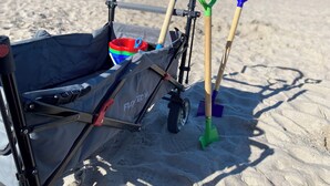 Ein Strandkorb mit schwarzem Bezug, einem roten und blauen Eimer sowie drei Schaufeln.
