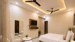 A modern hotel room with a bed, two chairs, a small table, a TV, and ceiling fans.