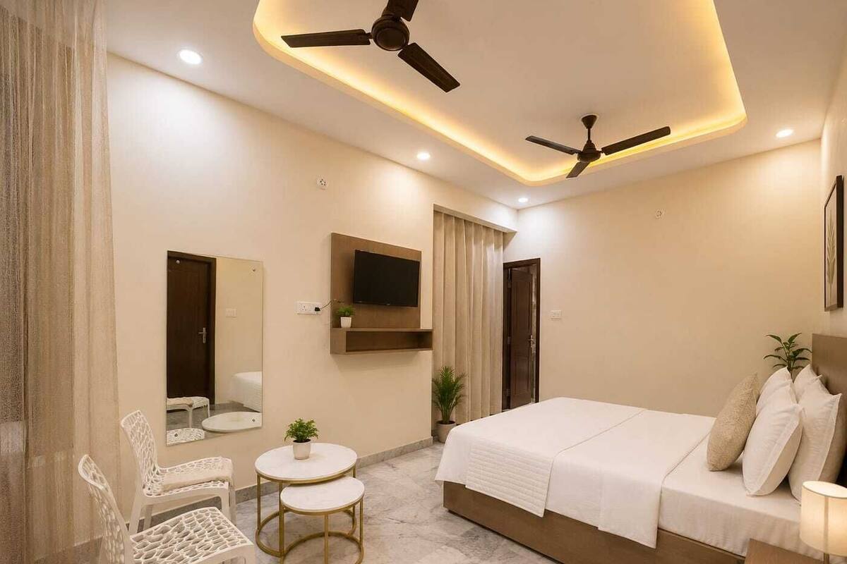 Ein modernes Hotelzimmer mit Bett, zwei Stühlen, einem kleinen Tisch, einem Fernseher und Deckenventilatoren.