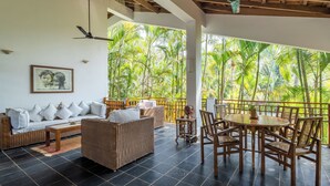 A room with a sofa, dining table, chairs, a ceiling fan, and a view of lush greenery. - Staymaster Ashvini 2BR Villa Jet Pool (Nerul)