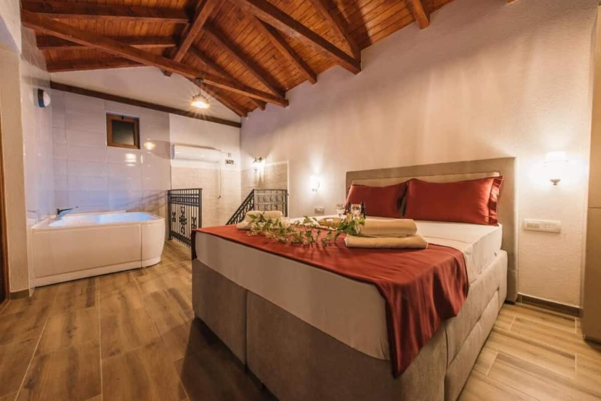 A bedroom with a bed, a red blanket, a wooden ceiling, a white wall, and a bathroom with a bathtub.
